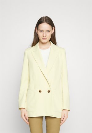 Jauna moteris vilki šviesiai geltoną dvigubą švarką su želvos skydo sagomis ir smėlio spalvos kelnėmis, stovinti prieš paprastą baltą foną.