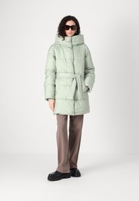 Lys grønn puffjakke med belte i midjen, høy krage og stor hette; har en quiltet design og er parret med brune bukser og svarte sko.