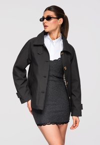 Femme aux cheveux bruns attachés en queue de cheval, portant des lunettes de soleil noires, une veste noire boutonnée sur une chemise blanche et une jupe rayée avec un bord en dentelle.