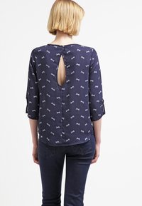 Blouse marine avec un motif de nœud, manches trois-quarts et dos en forme de trou de serrure. Texture lisse, tissu léger. Associée à un jean foncé.