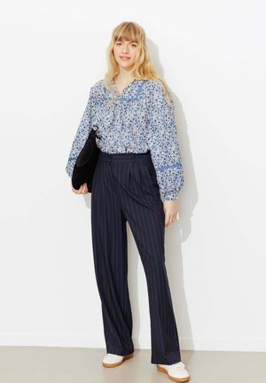 Jeune femme portant un chemisier bleu à fleurs, un pantalon large à rayures bleu marine, des baskets blanches, tenant un sac noir contre un mur blanc.