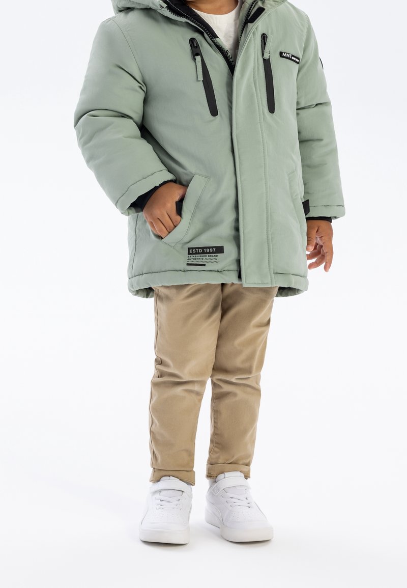 Un enfant debout portant une veste d'hiver vert clair avec des fermetures éclair noires, un pantalon beige et des baskets blanches sur un fond blanc.