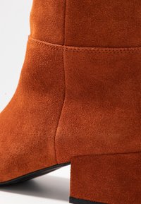 Botín de ante en color naranja quemado con punta cuadrada y tacón bajo en bloque, que presenta una textura suave y un detalle de costura en contraste.