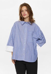 Blue and white striped button-up shirt with a chest pocket, featuring wide sleeves with white cuffs and a relaxed fit. Soft fabric texture.