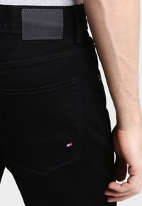 Zbliżenie na talię osoby noszącej czarne dżinsy Tommy Hilfiger z haftowanym tylnym kieszonką i skórzaną metką marki na pasku.