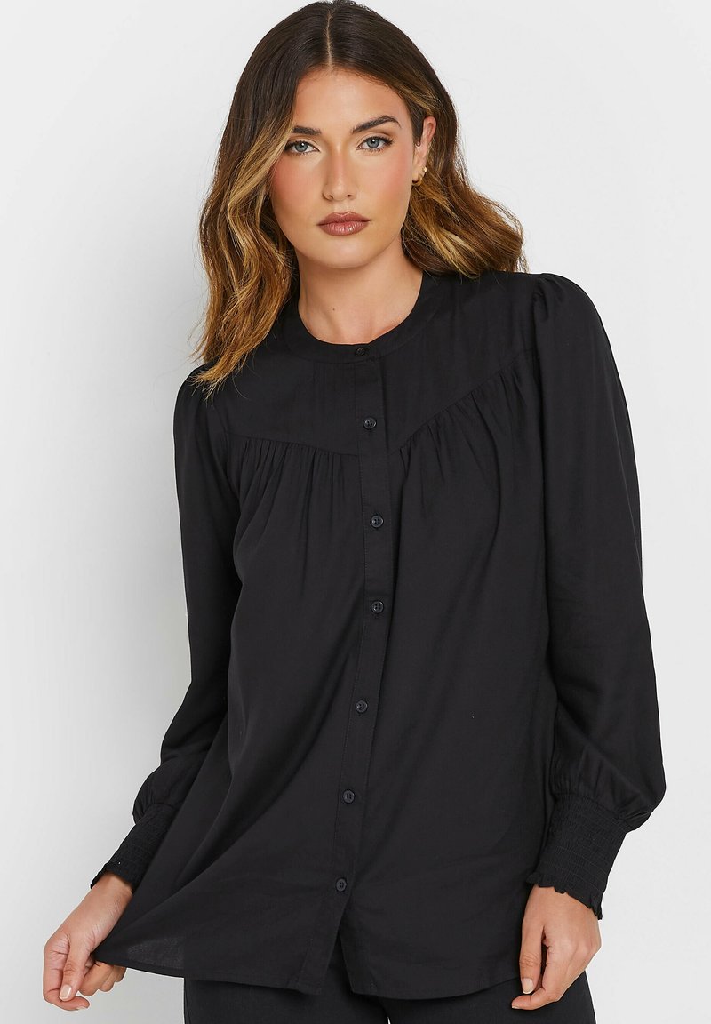 Camisa negra de manga larga con tela recogida sobre el pecho, cierre de botones y puños fruncidos. Material suave y ajuste holgado.