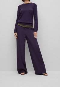 Lila långärmad topp med en metallisk accent vid midjan, kombinerad med vida lila byxor och sandaletter med klack.