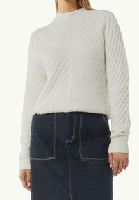 Pull en tricot blanc avec un col montant côtelé, présentant des motifs texturés verticaux. Le mannequin porte une jupe en denim avec des coutures visibles.
