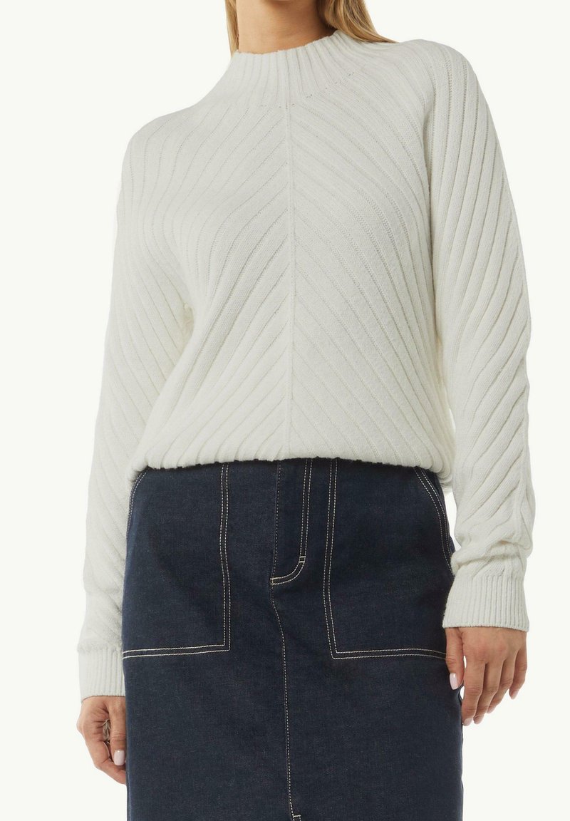 Pull en tricot blanc avec un col montant côtelé, présentant des motifs texturés verticaux. Le mannequin porte une jupe en denim avec des coutures visibles.