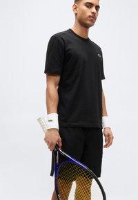 Νεαρός άνδρας με μαύρη αθλητική ενδυμασία κρατώντας ρακέτα τένις με λευκά περιβραχιόνια, στέκεται μπροστά σε απλό φωτεινό φόντο.