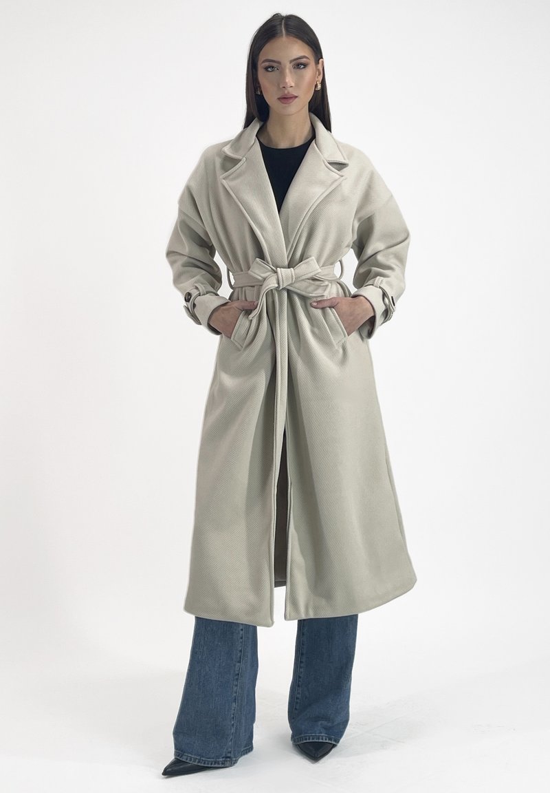 Cappotto beige chiaro con cintura, colletto a punta, grandi tasche e tessuto texture, abbinato a jeans blu a campana e scarpe nere a punta.