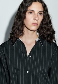 Persona con lunghi capelli ricci e scuri indossa una camicia nera con sottili righe verticali bianche, guardando leggermente verso destra su uno sfondo semplice.