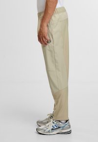 Beżowe spodenki sportowe z elastycznym pasem, bocznymi kieszeniami i logo, połączone z srebrno-niebieskimi butami do biegania. Gładka faktura.