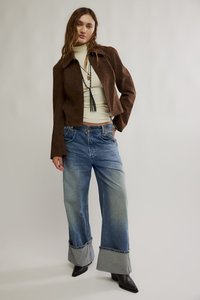 Braune Wildlederjacke in Cropped-Form mit weitem Kragen, cremefarbener Rollkragenpullover, blaue Jeans mit ausgestellten Beinen und umgeschlagenen Säumen sowie schwarze spitze Stiefel.