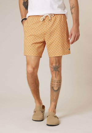 Homme portant un short orange à motifs avec un cordon blanc, des chaussures beige à enfiler, une chemise blanche, des bracelets et des tatouages visibles sur les jambes et le bras.
