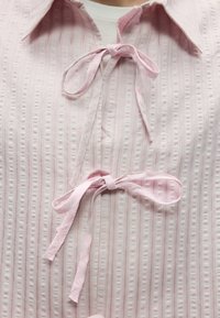 Ροζ τοπ με υφή, κάθετες ραβδώσεις, γιακά και μπροστινά κορδόνια με κορδέλες. Το υλικό φαίνεται ελαφρύ και απαλό.