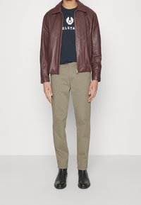 Man med burgundy läderjacka, marinblå tryckt t-shirt, beige byxor och svarta läderankelstövlar som står mot vit bakgrund.