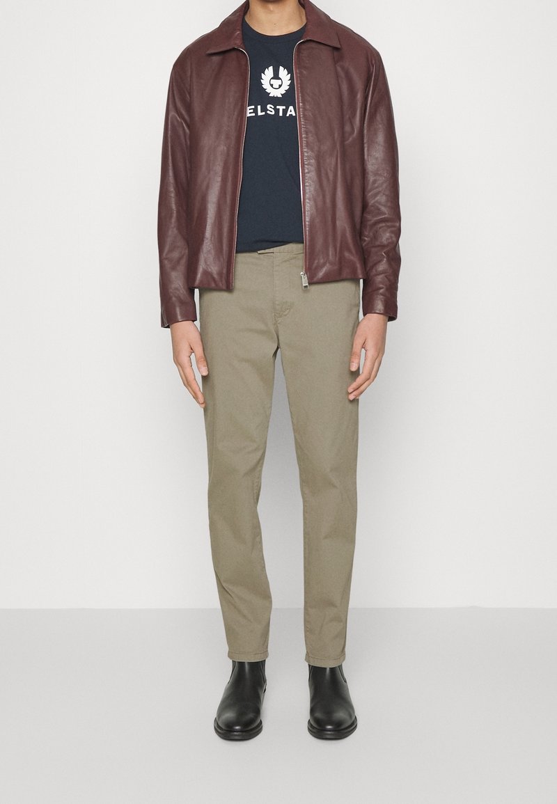 Man med burgundy läderjacka, marinblå tryckt t-shirt, beige byxor och svarta läderankelstövlar som står mot vit bakgrund.