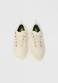 Αθλητικά παπούτσια ανοιχτό μπεζ με υφή διχτυού στο πάνω μέρος, στρογγυλεμένη μύτη και αντιθετικές μαύρες σόλες με μια μικρή πράσινη ετικέτα.