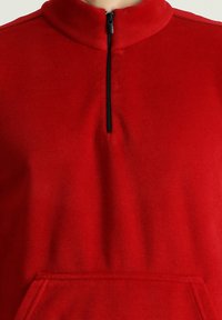 Suéter rojo hecho de tela de forro polar, con un cuello de media cremallera y un bolsillo frontal. Textura suave con detalle de cremallera negra.