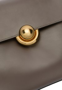 Pochette en cuir gris avec un fermoir circulaire en or. Texture lisse, matériel centré avec le nom de la marque gravé, et bords surpiqués.