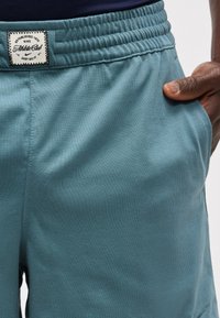 Κοντινό πλάνο σε πετρόλ αθλητικό σορτς με ελαστική μέση και ετικέτα Nike Athletic Club, που φοριέται από άτομο με το χέρι στην τσέπη.