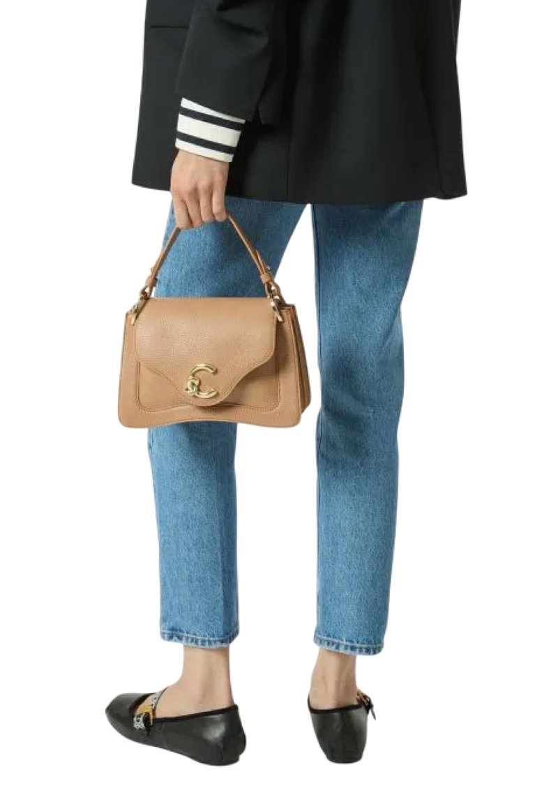 Beige textuur handtas met gouden accessoires, vastgehouden door een persoon in een zwarte blazer en blauwe jeans, gedragen met zwarte ballerina's.