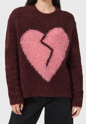 Femme portant un pull duveteux bordeaux avec un grand motif de cœur brisé rose à l'avant, associé à un pantalon noir.