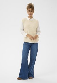 Bež pleteni bezrukavnik preko bijele košulje s ovratnikom, u kombinaciji s raširenim plavim trapericama i smešim obućom, na bijeloj pozadini.