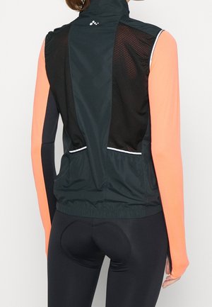 Woman wearing black sleeveless cycling vest with mesh back panels over long-sleeve neon orange and black top, and black padded cycling shorts.