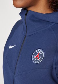 Person, die eine blaue Paris Saint-Germain-Jacke mit einem Reißverschlusskragen und einem weißen Nike-Swoosh-Logo auf der Brust trägt.