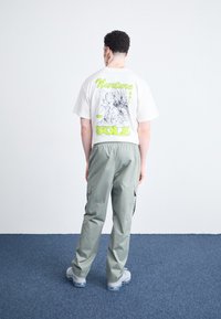 Γραφικό μπλουζάκι σε εκρού, με πράσινο κείμενο και σχέδιο στην πλάτη. Συνδυασμένο με ανοιχτά πράσινα cargo παντελόνια και γκρι αθλητικά παπούτσια.