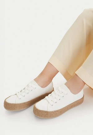 Sneakers bianche con lacci e suole spesse in juta intrecciata, indossate con pantaloni beige chiaro che lasciano scoperta la caviglia, su sfondo semplice.