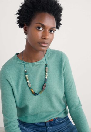 Pull en tricot vert clair avec col rond, associé à un jean bleu. Un collier en perles multicolores avec diverses textures et designs pend autour du cou.