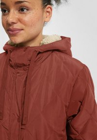 Braune gesteppte Jacke mit Kapuze, mit einem weichen beige Fleece-Futter, Zugbändern und Druckknöpfen auf der Vorderseite.