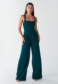 Teal jumpsuit with wide legs, square neckline, and thick straps. Made of smooth fabric, featuring pleats at the waist and minimalistic design.
