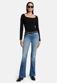 Zwarte lange mouwen top, blauwe flared jeans met versleten details, zwarte riem en zwarte laarzen, gecombineerd met hoekige zwarte zonnebril.