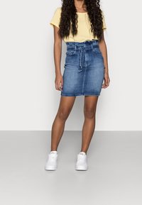 Jeanskjol med en figurnära silhuett och bälte runt midjan, kombinerad med en gul kortärmad topp och vita sneakers mot en grå bakgrund.