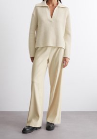 Pull en maille côtelée beige avec col en V et large col, associé à un pantalon large beige clair, et des mocassins à plateforme noirs.