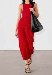 Czerwona sukienka bez rękawów z węzłem w talii, zestawiona z płaskimi czarnymi sandałami i dużą czarną torbą. Gładki materiał, długość do ziemi.