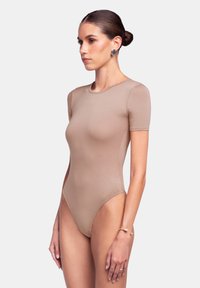 Body a maniche corte, di colore nude, con una forma aderente e una texture liscia, caratterizzato da un collo rotondo e gambe in alto per un aspetto affusolato.