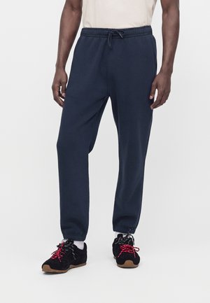 Hombre con pantalones de chándal azul marino con cintura elástica y cordón, combinados con zapatillas negras con cordones rojos y calcetines blancos sobre un fondo blanco.