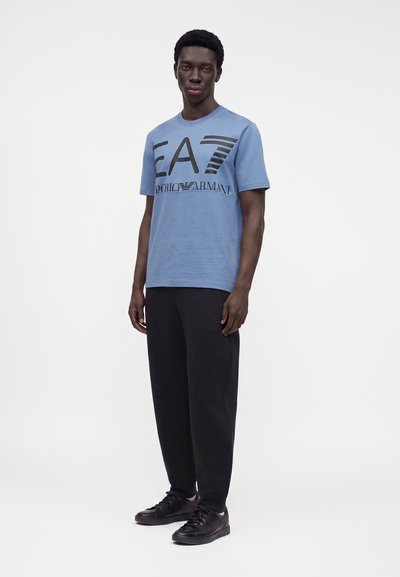 Νέος άνδρας με μπλε μπλουζάκι EA7 Emporio Armani, μαύρο παντελόνι και μαύρα αθλητικά παπούτσια, στέκεται μπροστά σε ένα απλό φωτεινό φόντο.