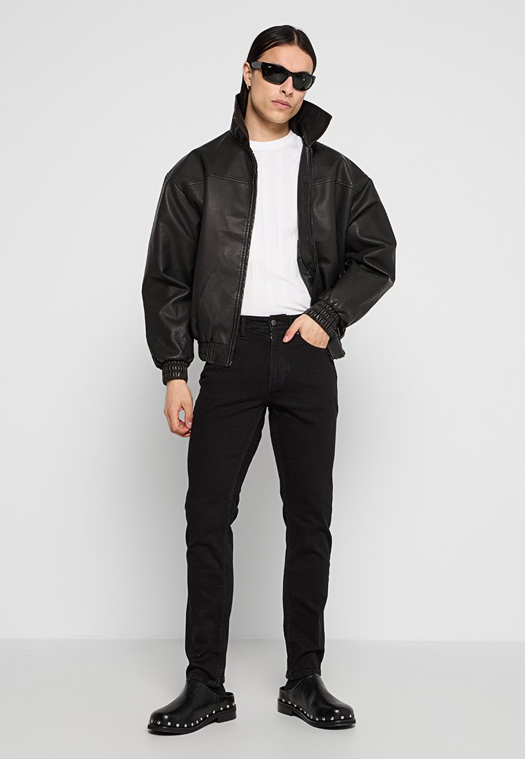 Veste bomber en cuir noir avec un col haut, chemise blanche en dessous, jeans skinny noirs, chaussures noires à clous et lunettes de soleil noires.