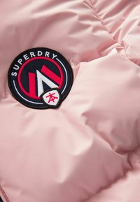Šviesiai rožinė pūkinė striukė, pagaminta iš audinio, su juodu apvaliu užsiuvimu, kuriame yra raudonos ir baltos spalvos akcentų, rodančių "Superdry" logotipą ir dizaino elementus.