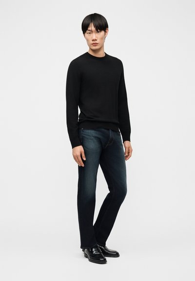 Νέος άντρας στέκεται προς τα εμπρός φορώντας μαύρο πουλόβερ με μακριά μανίκια, σκούρο μπλε τζιν και μαύρα δερμάτινα παπούτσια μπροστά σε απλό φόντο.