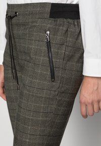 Grå rutiga byxor med svart elastisk midja och dragsko, med en vertikal svart dragkedjeficka på högra sidan, bärs med en vit skjorta.