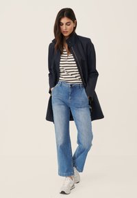 Marineblå letvægtsjakke med høj krave, stribet t-shirt, lyseblå brede jeans og grå sneakers. Modellen poser med hænderne i lommerne.