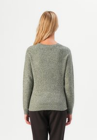 Woman with long blonde hair stands facing away, wearing a light gray knit sweater and dark pants against a plain white background.