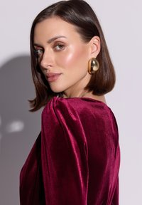 Bordowa aksamitna bluzka z bufkami na rękawach. Modelka ma gładkie włosy ścięte na boba i nosi duże złote kolczyki koła. Delikatne oświetlenie podkreśla fakturę.